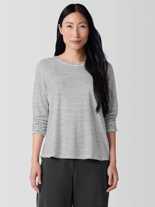 A person with long black hair wearing an Eileen Fisher Striped Organic Linen Jersey Long-Sleeve Tee in White/Black and dark pants smiles slightly, standing against a plain gray background.