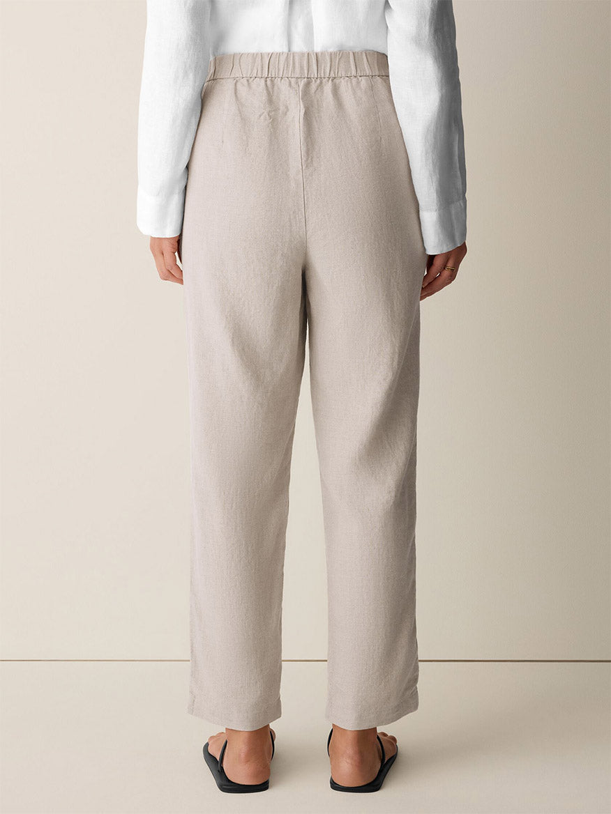 Person standing with their back to the camera, wearing the Eileen Fisher Organic Linen Tapered Pant in Undyed Natural, paired with a white long-sleeve shirt and black sandals against a plain background.