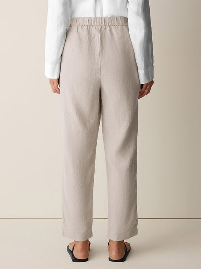 Person standing with their back to the camera, wearing the Eileen Fisher Organic Linen Tapered Pant in Undyed Natural, paired with a white long-sleeve shirt and black sandals against a plain background.