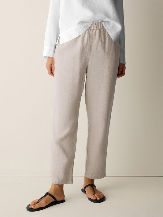 A person wears the Eileen Fisher Organic Linen Tapered Pant in Undyed Natural with a white long-sleeve shirt and black sandals, standing against a plain background. The pants feature an elastic waistband and ankle-length tapered fit.