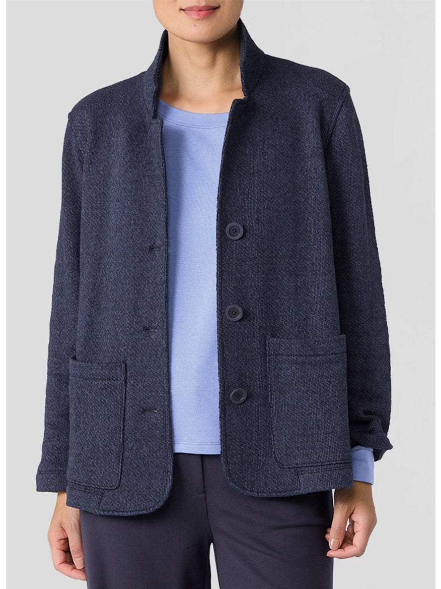 Standing against a light gray background, the person is dressed in the Eileen Fisher Organic Cotton Terry Stand Collar Jacket in Ink. This dark blue jacket is made from organic cotton and features patch pockets, beautifully paired with a light blue shirt underneath.