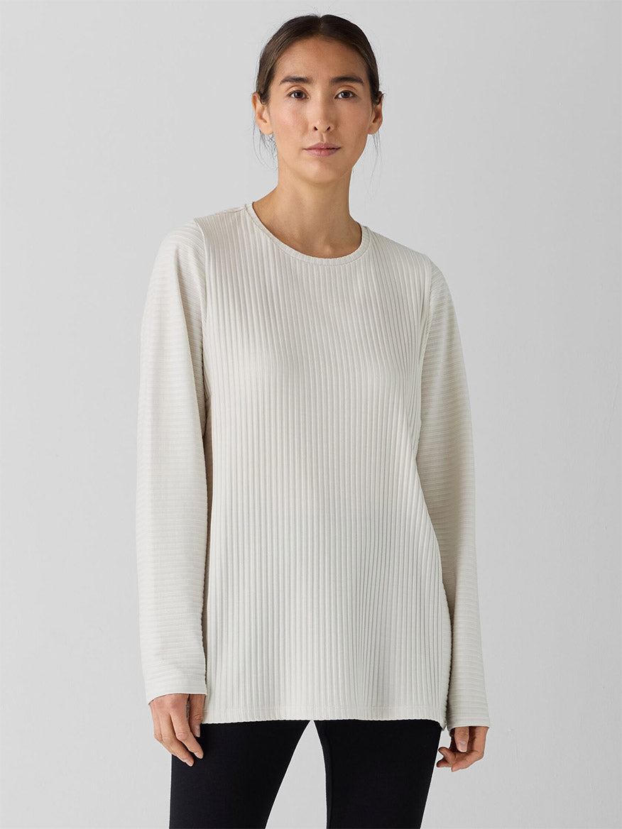 Wearing the Eileen Fisher Textured Stretch Rib Round Neck Long-Sleeve Top in Bone and black pants, a person stands against a plain backdrop, highlighting the elegant ribbed texture of their attire.