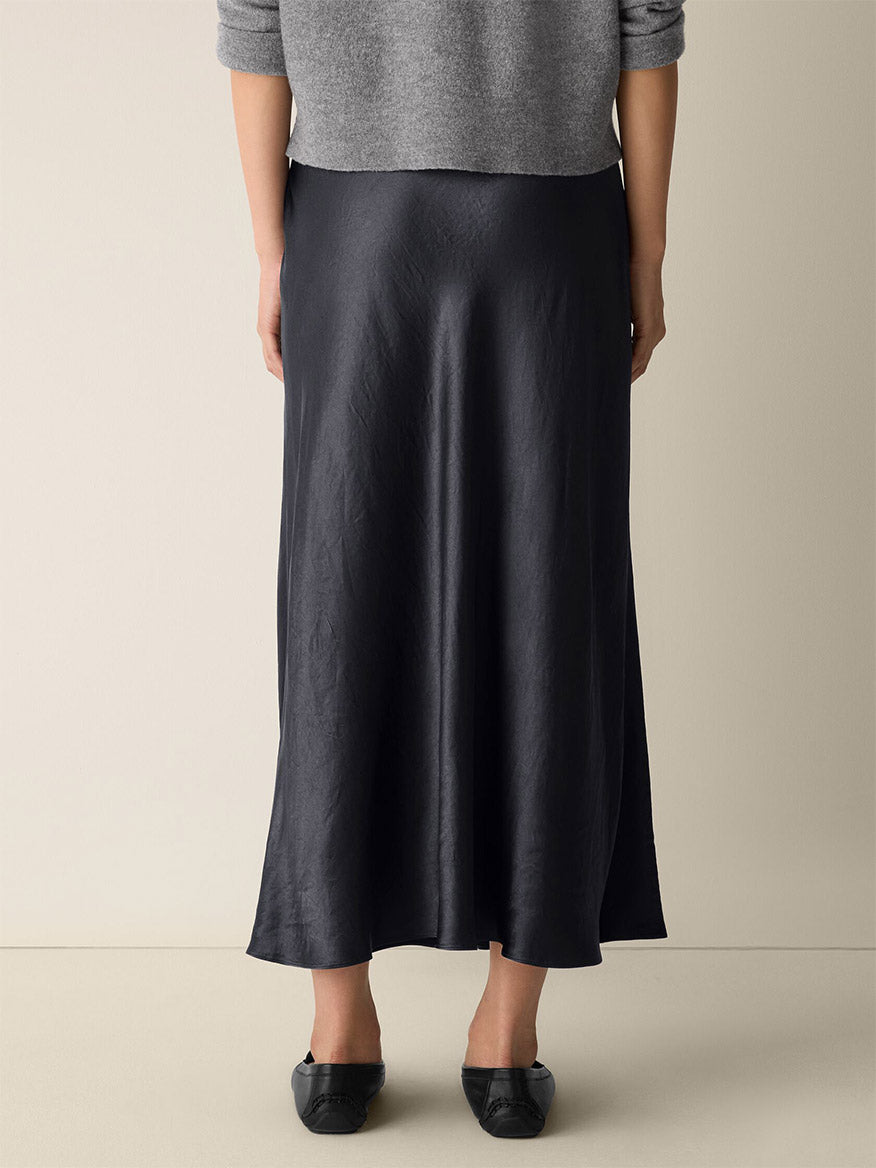 A person stands facing away, dressed in a grey sweater, black flat shoes, and the Eileen Fisher Textured Satin Slip Skirt in Graphite featuring an A-line silhouette.