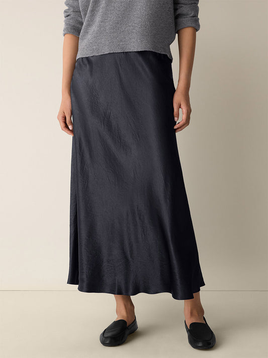 Person wearing a grey long-sleeve sweater, Eileen Fisher Textured Satin Slip Skirt in Graphite, and black slip-on shoes, standing against a plain beige background.