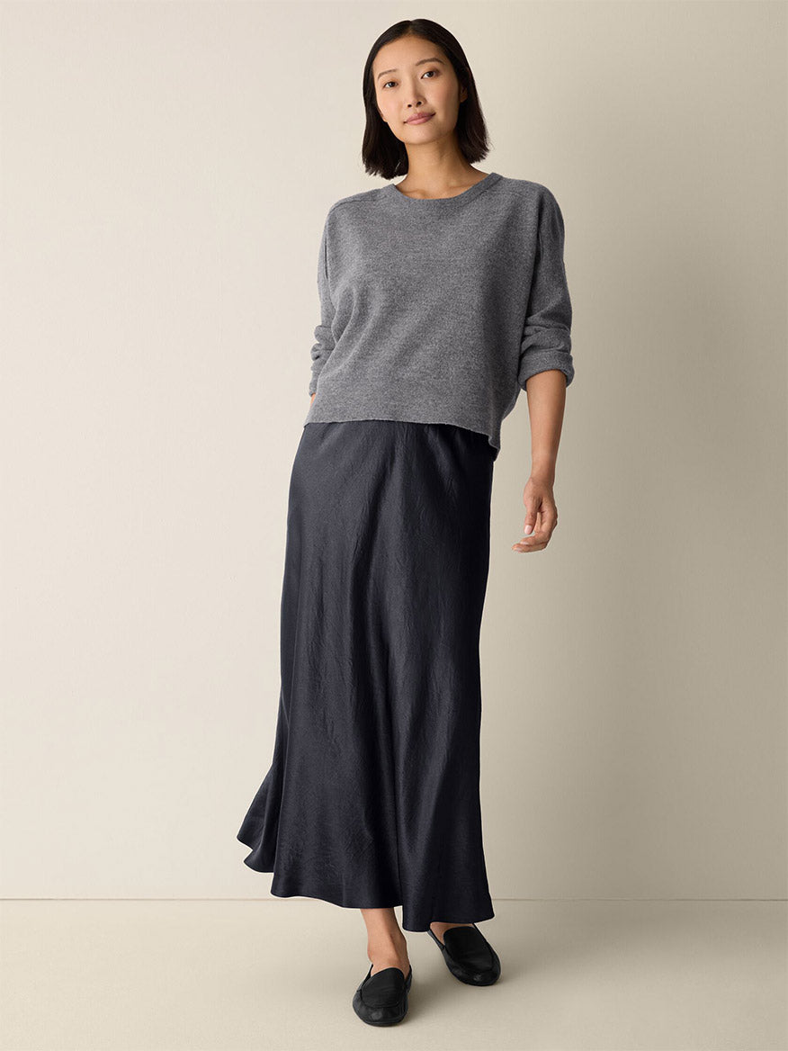 A woman stands against a plain background wearing a grey sweater, black flat shoes, and the Eileen Fisher Textured Satin Slip Skirt in Graphite.