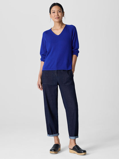 A person stands against a plain background wearing an Eileen Fisher Peruvian Cotton Blend V-Neck Top in Lapis, dark rolled-up jeans, and black slip-on shoes, with hands in pockets.