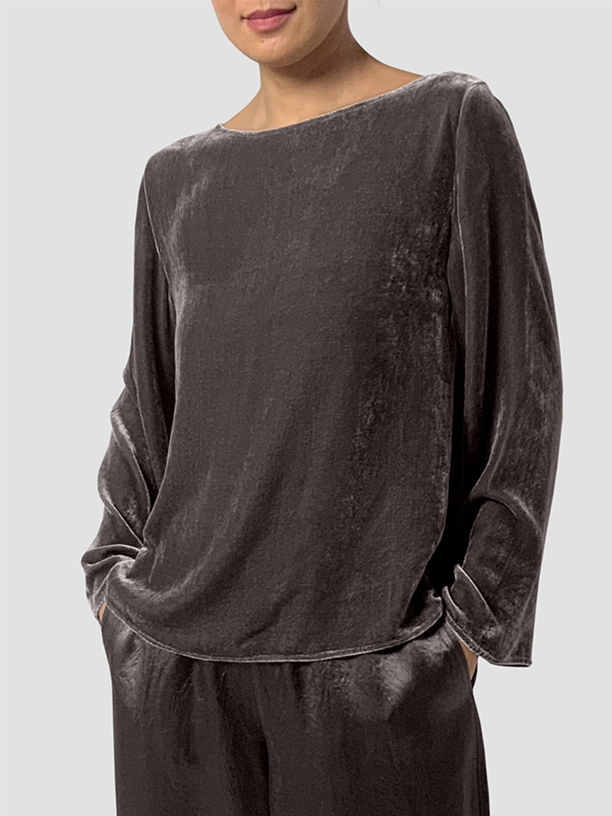 Someone wearing the Eileen Fisher Velvet Bateau Neck Top in Rye, a loose-fitting, long-sleeved, dark grey Fair Trade Certified top with matching pants, stands with hands in pockets against a plain background.