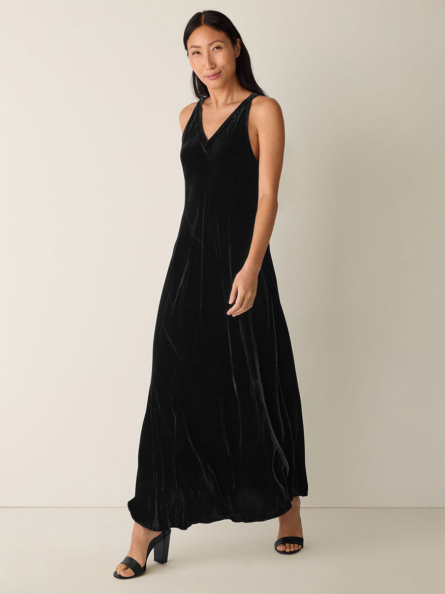 A woman stands against a plain background wearing the Eileen Fisher Velvet V-Neck Dress in Black and black heeled sandals.