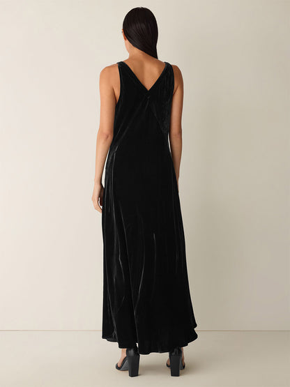 A person with long dark hair stands with their back to the camera against a plain light background, wearing the Eileen Fisher Velvet V-Neck Dress in Black and black heels.