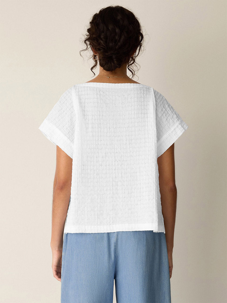 A person with curly hair, facing away, wears the Eileen Fisher Organic Cotton Voile Box Square Top in white and light blue pants against a plain background.