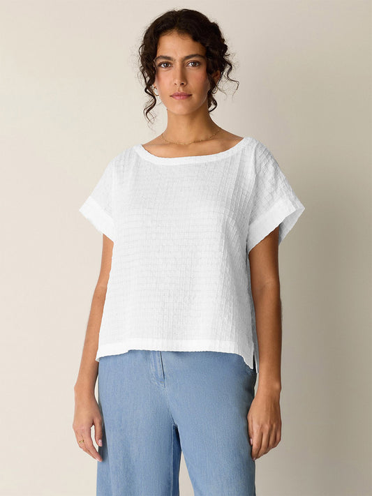 A woman stands against a plain light background, wearing the Eileen Fisher Organic Cotton Voile Box Square Top in white and light blue jeans, facing forward with a neutral expression.