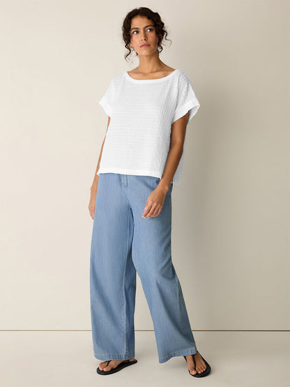 A woman stands against a plain wall wearing the Eileen Fisher Organic Cotton Voile Box Square Top in White, light blue wide-leg jeans, and black sandals.