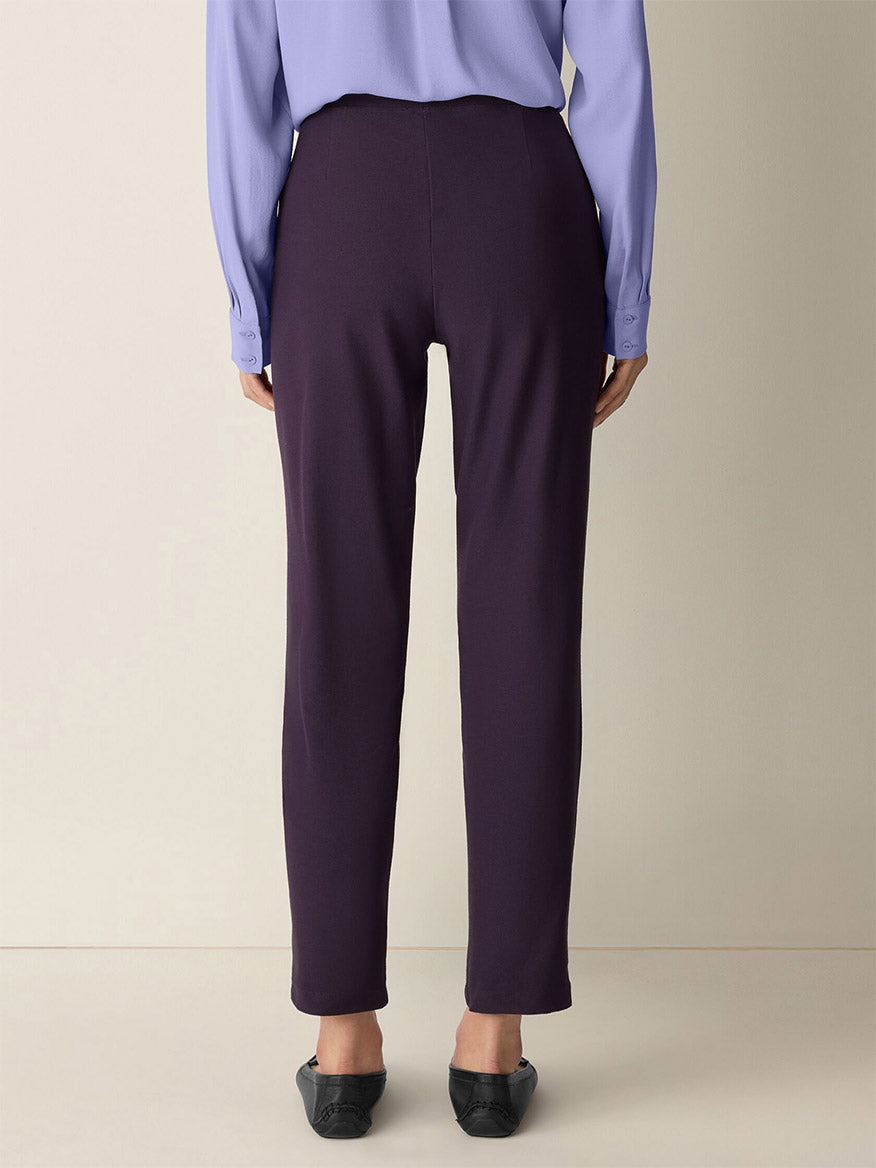 A person stands facing away, wearing a light purple blouse, black flats, and the Eileen Fisher Washable Stretch Crepe Pant in Aubergine—perfect for stylish comfort with easy-care convenience.