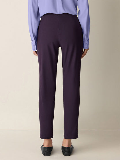 A person stands facing away, wearing a light purple blouse, black flats, and the Eileen Fisher Washable Stretch Crepe Pant in Aubergine—perfect for stylish comfort with easy-care convenience.