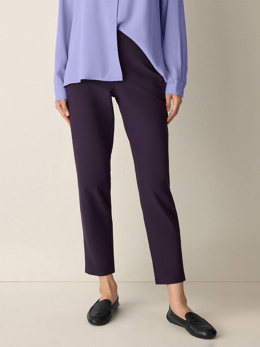 A person wearing a light purple blouse, Eileen Fisher Washable Stretch Crepe Pant in Aubergine, and black loafers stands against a beige background.