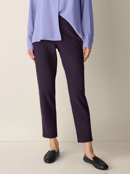A person wearing a light purple blouse, Eileen Fisher Washable Stretch Crepe Pant in Aubergine, and black loafers stands against a beige background.