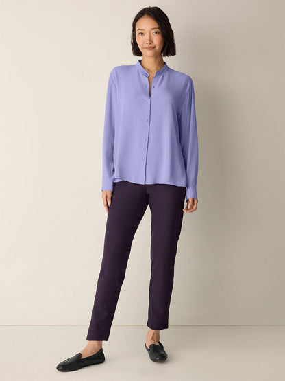 A woman stands against a plain wall wearing a light purple button-up shirt, black flats, and the Eileen Fisher Washable Stretch Crepe Pant in Aubergine for an effortlessly stylish, washable look.