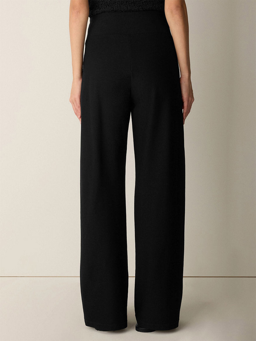 A person stands facing away against a plain light background, wearing the Eileen Fisher Washable Stretch Crepe Pintuck Wide Pant in Black paired with a black top. Only the lower back and legs are visible, highlighting the pant’s elegant drape and showcasing its easy-care, washable stretch crepe fabric.