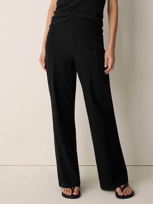 A person wearing Eileen Fisher Washable Stretch Crepe Pintuck Wide Pant in Black, a black top, and black sandals stands against a plain off-white background.
