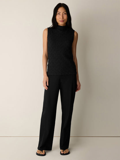 A woman stands against a plain wall wearing a sleeveless black textured top, the Eileen Fisher Washable Stretch Crepe Pintuck Wide Pant in Black, and black sandals.