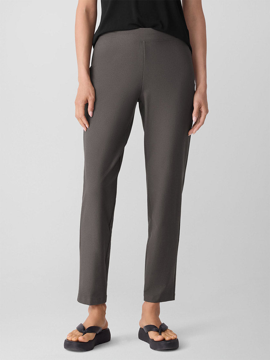 A person wearing gray Eileen Fisher Washable Stretch Crepe Pant in Rye, a black top, and black sandals stands against a plain light background.