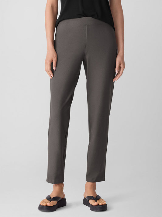 A person wearing gray Eileen Fisher Washable Stretch Crepe Pant in Rye, a black top, and black sandals stands against a plain light background.
