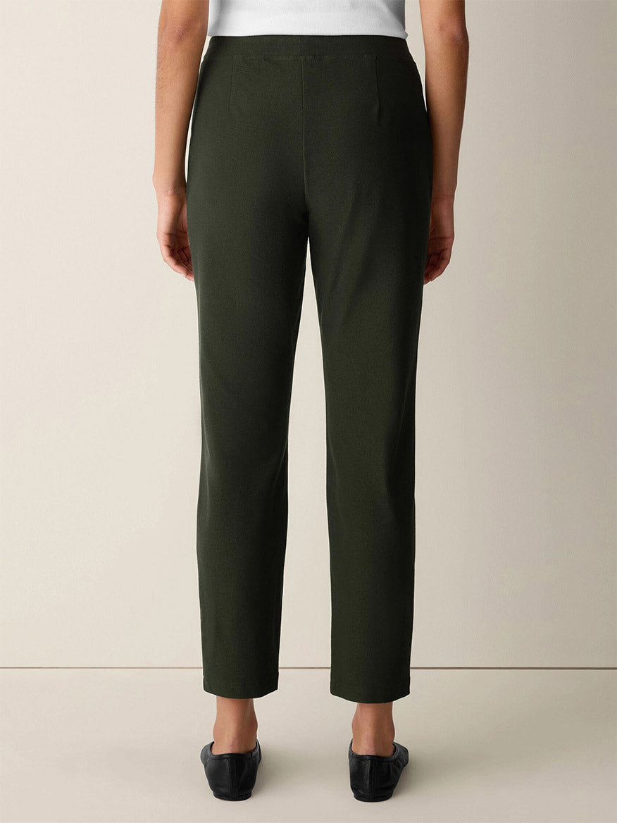 A person wearing the Eileen Fisher Washable Stretch Crepe Slim Pant in Seaweed and black shoes, standing against a plain light-colored background, shown from the back.