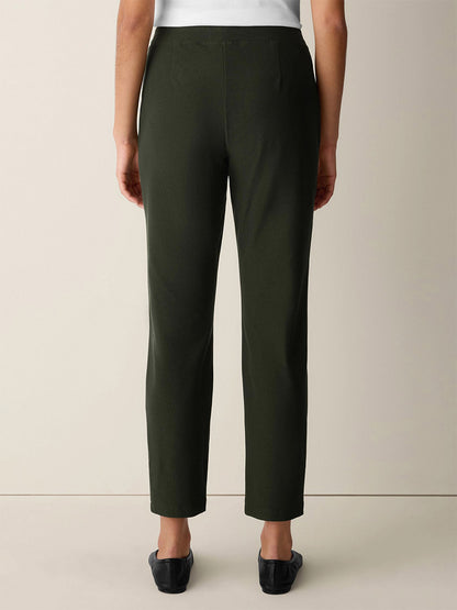 A person wearing the Eileen Fisher Washable Stretch Crepe Slim Pant in Seaweed and black shoes, standing against a plain light-colored background, shown from the back.