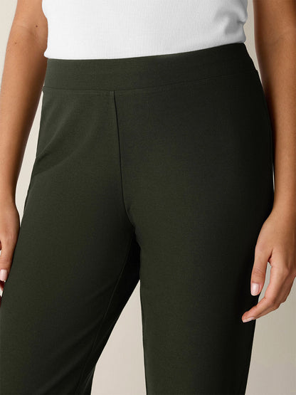 A person is shown from the waist to mid-thigh wearing the Eileen Fisher Washable Stretch Crepe Slim Pant in Seaweed—a deep green shade—paired with a white ribbed top, one arm relaxed at their side.