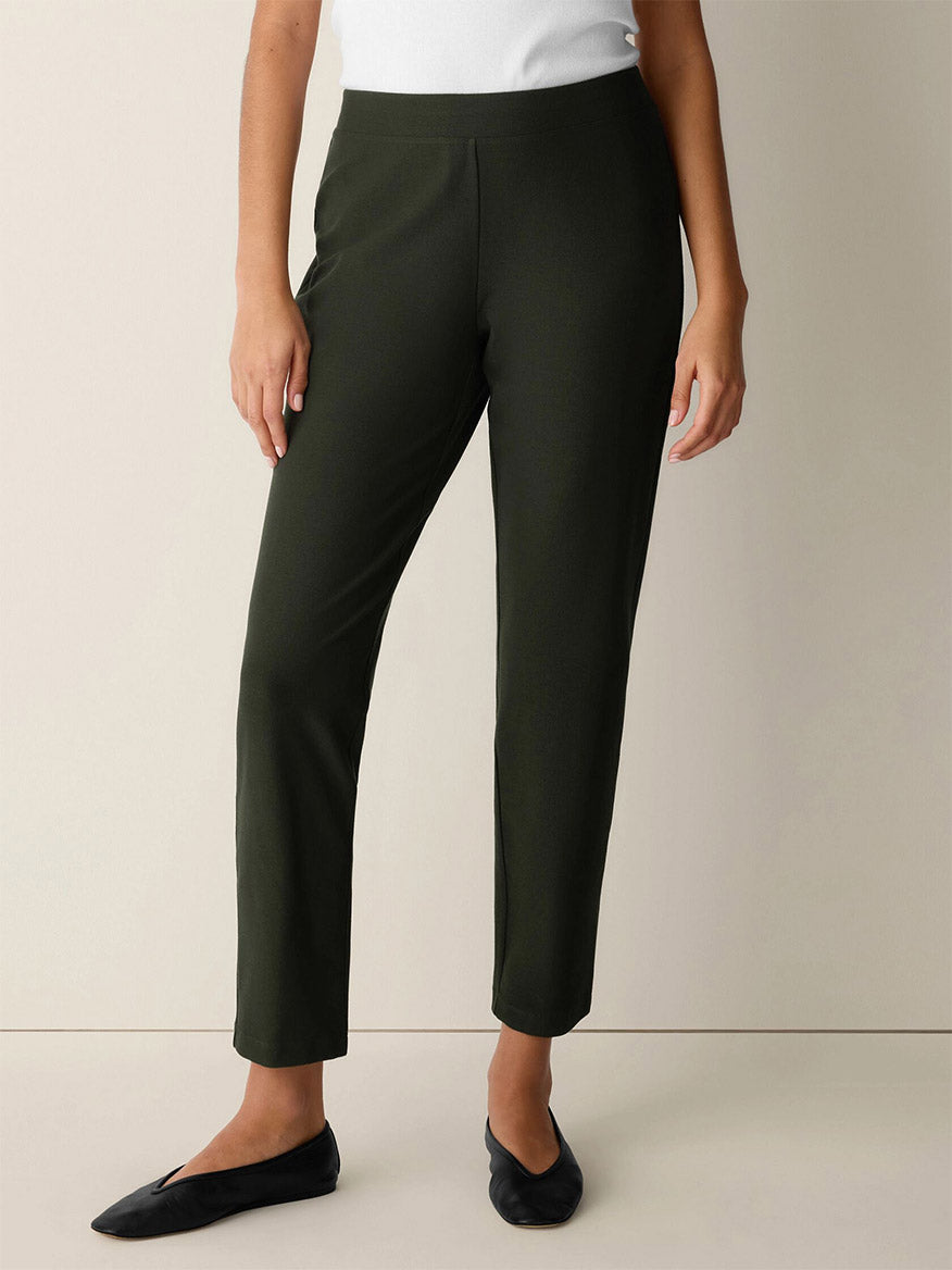Wearing the Eileen Fisher Washable Stretch Crepe Slim Pant in Seaweed, paired with a white top and black flats, the person stands against a plain light-colored background.