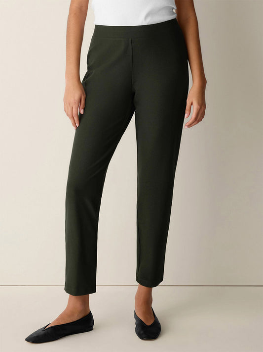 Wearing the Eileen Fisher Washable Stretch Crepe Slim Pant in Seaweed, paired with a white top and black flats, the person stands against a plain light-colored background.