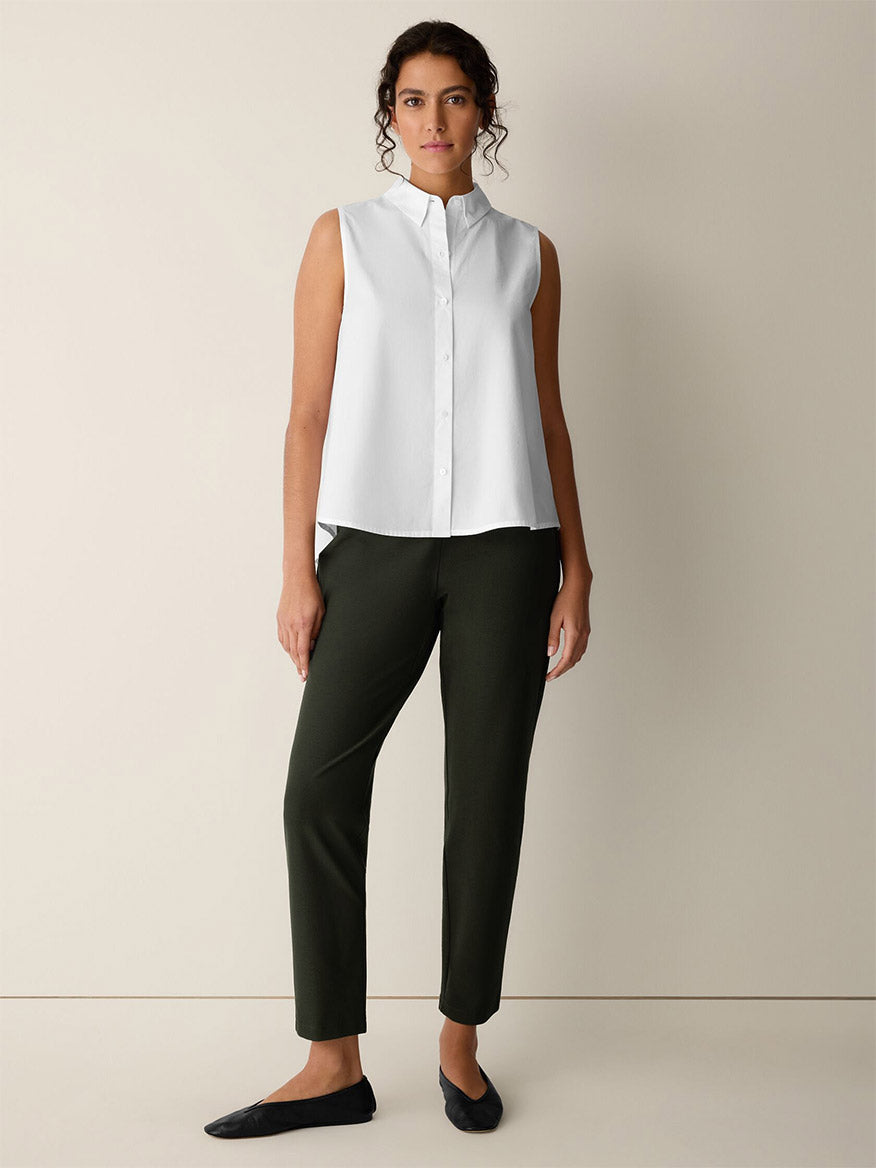 A woman stands against a plain wall wearing a sleeveless white button-up shirt, black flats, and Eileen Fisher Washable Stretch Crepe Slim Pants in Seaweed.