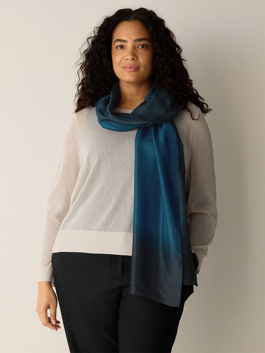 Woman with long curly hair wears a light sweater, black pants, and an elegant Eileen Fisher Watercolor Organic Silk Scarf in Sailor Blue, standing against a plain light background.