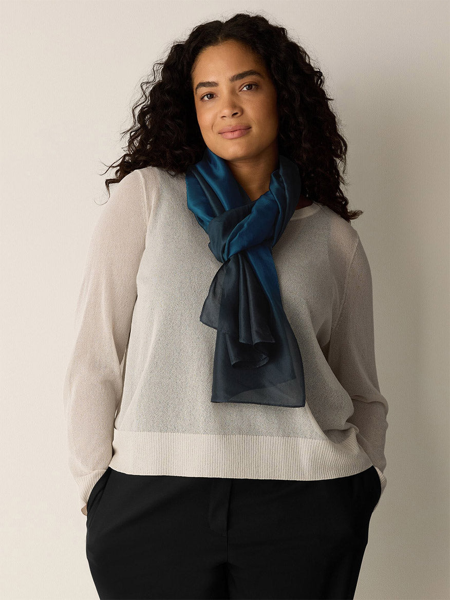 A woman with curly hair wears a sheer white sweater, black pants, and the Eileen Fisher Watercolor Organic Silk Scarf in Sailor Blue—a fair trade accessory featuring a blue gradient—as she stands against a plain light background with her hands in her pockets.