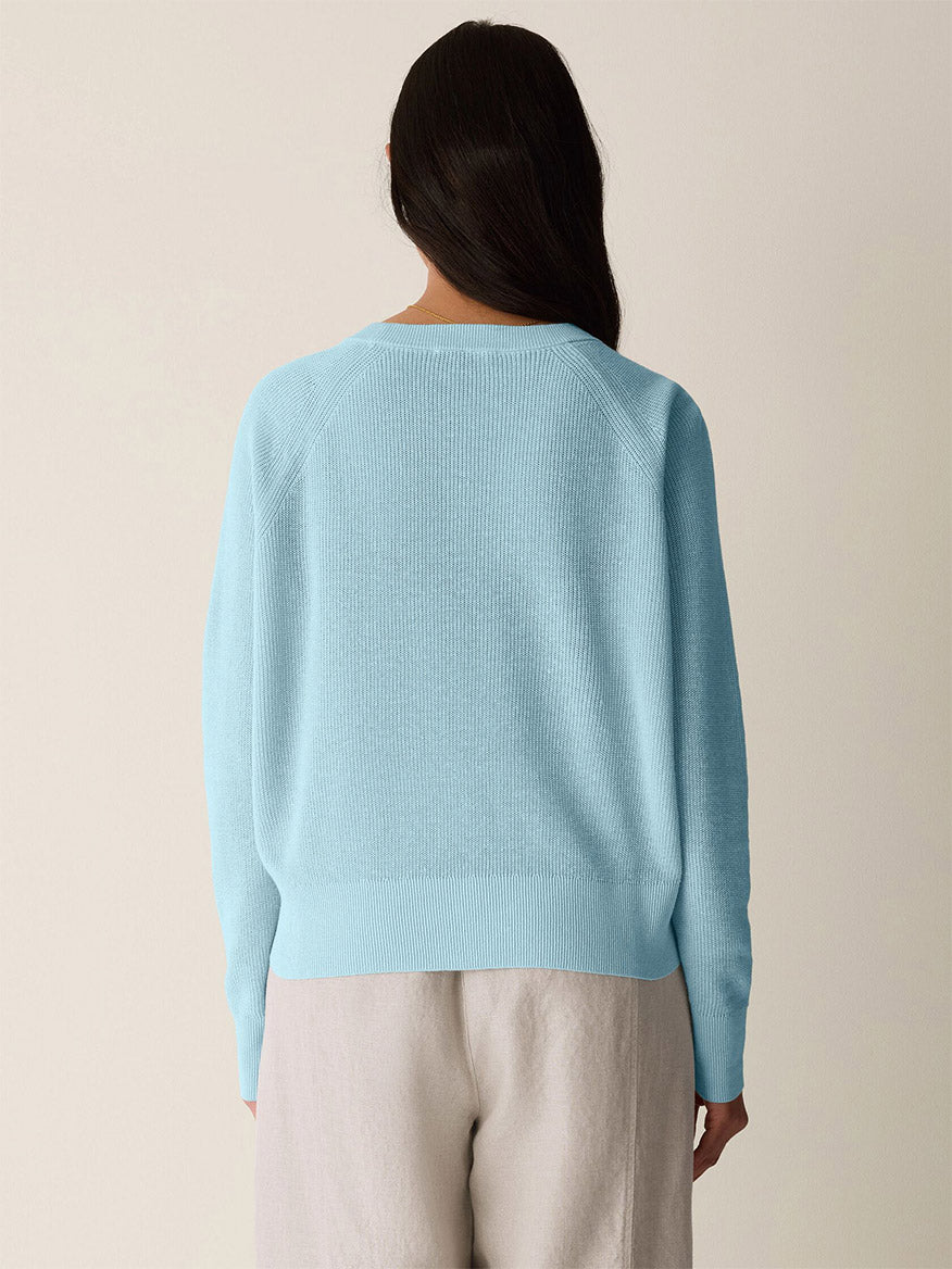 A person with long dark hair is seen from the back, wearing the Eileen Fisher Organic Linen Cotton Wedge Sweater in Breeze and beige pants, standing against a plain light background.