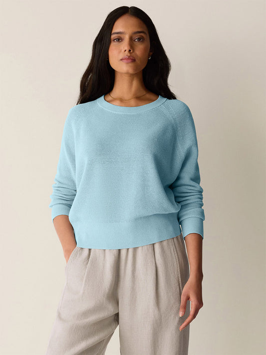 A woman stands facing forward, hands in her pockets, wearing the Eileen Fisher Organic Linen Cotton Wedge Sweater in Breeze and beige pants against a plain background.