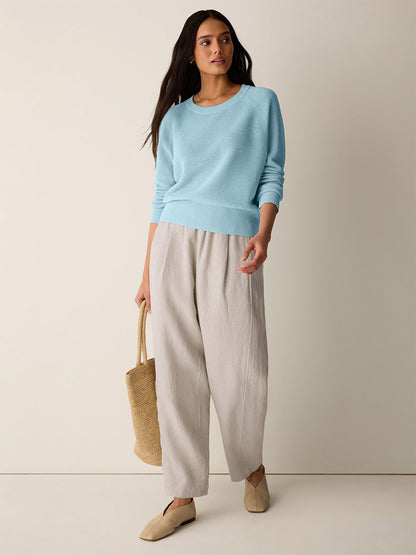 A woman stands against a plain background wearing the Eileen Fisher Organic Linen Cotton Wedge Sweater in Breeze, paired with beige wide-leg pants, beige shoes, and holding a woven tote bag.