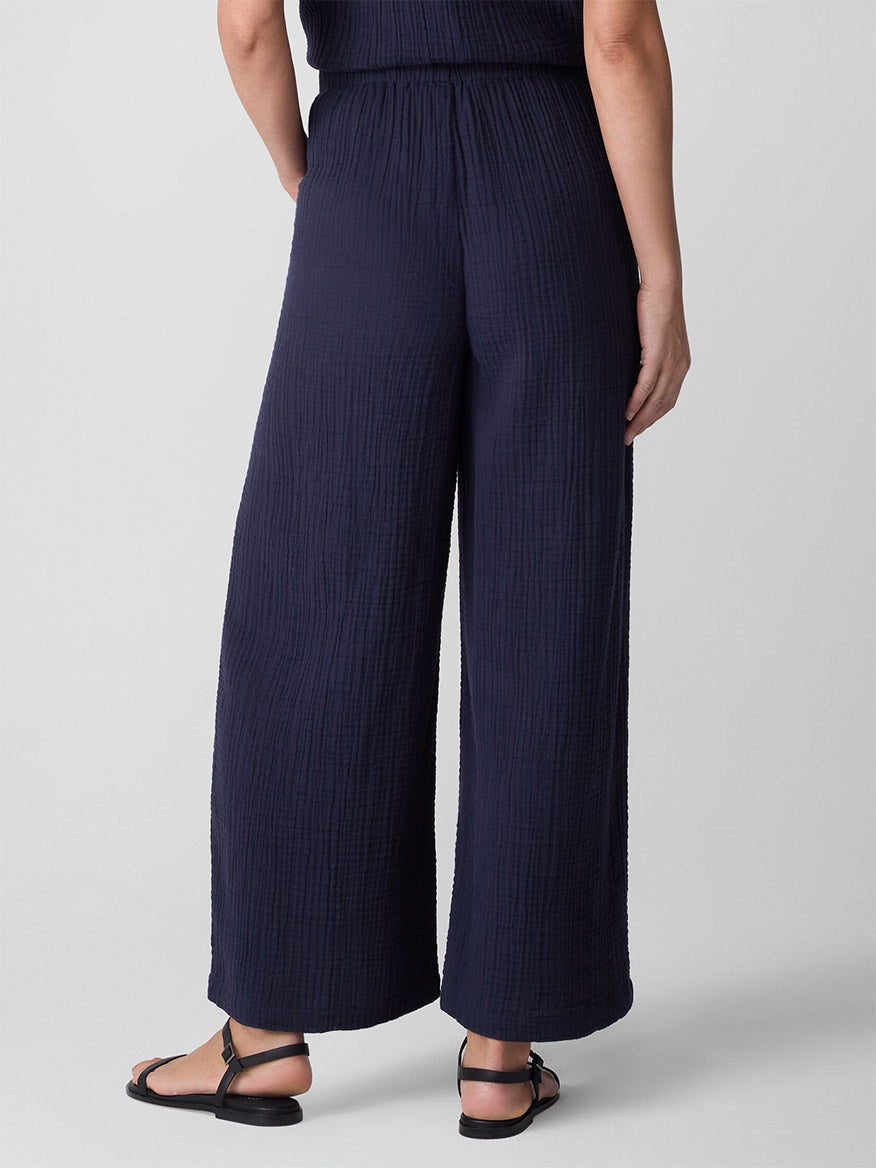 Wearing the Eileen Fisher Organic Cotton Lofty Gauze Wide-Leg Pant in Ink, styled with black sandals, a person stands against a plain light background, shown from the back.