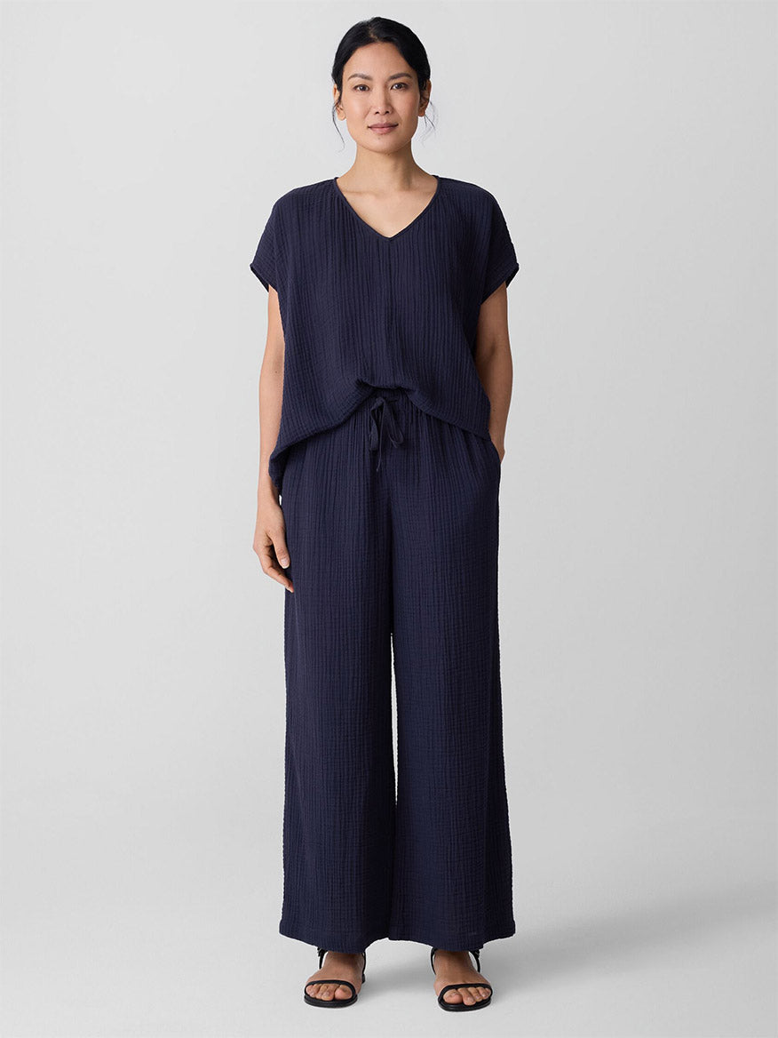 A woman stands facing forward, wearing the Eileen Fisher Organic Cotton Lofty Gauze Wide-Leg Pant in Ink with a matching navy top and sandals, set against a plain light gray background.