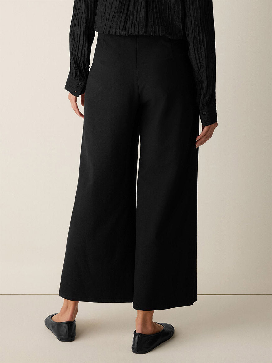 A person indoors is shown from the back wearing black flats, a long-sleeve textured blouse, and Eileen Fisher Washable Stretch Crepe Wide-Leg Pant in Black with an elastic waist.