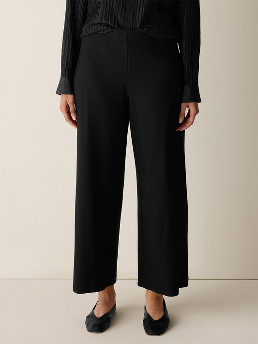 A person wearing Eileen Fisher Washable Stretch Crepe Wide-Leg Pant in black, a black long-sleeve shirt, and black flat shoes stands against a plain light background.