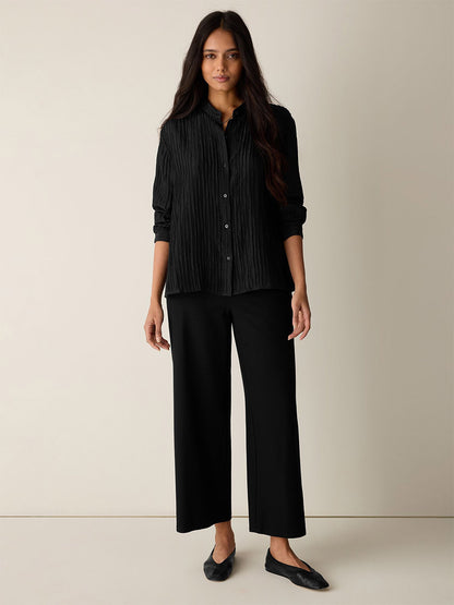 A woman stands against a plain light wall, wearing a black button-up pleated shirt, black flats, and Eileen Fisher Washable Stretch Crepe Wide-Leg Pant in Black with an elastic waist.