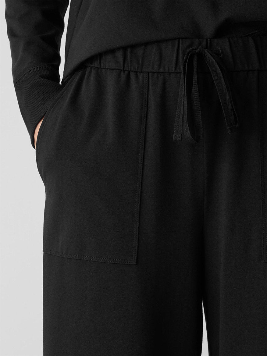 Close-up of a person wearing Eileen Fisher Pima Cotton Stretch Jersey Wide-Leg Pant in black, featuring a relaxed fit, drawstring waist, large front pockets, and one hand in a pocket.