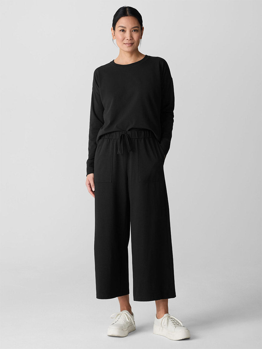 A woman stands against a plain background, wearing a black long-sleeve top and Eileen Fisher Pima Cotton Stretch Jersey Wide-Leg Pant in Black with pockets, paired with white sneakers.