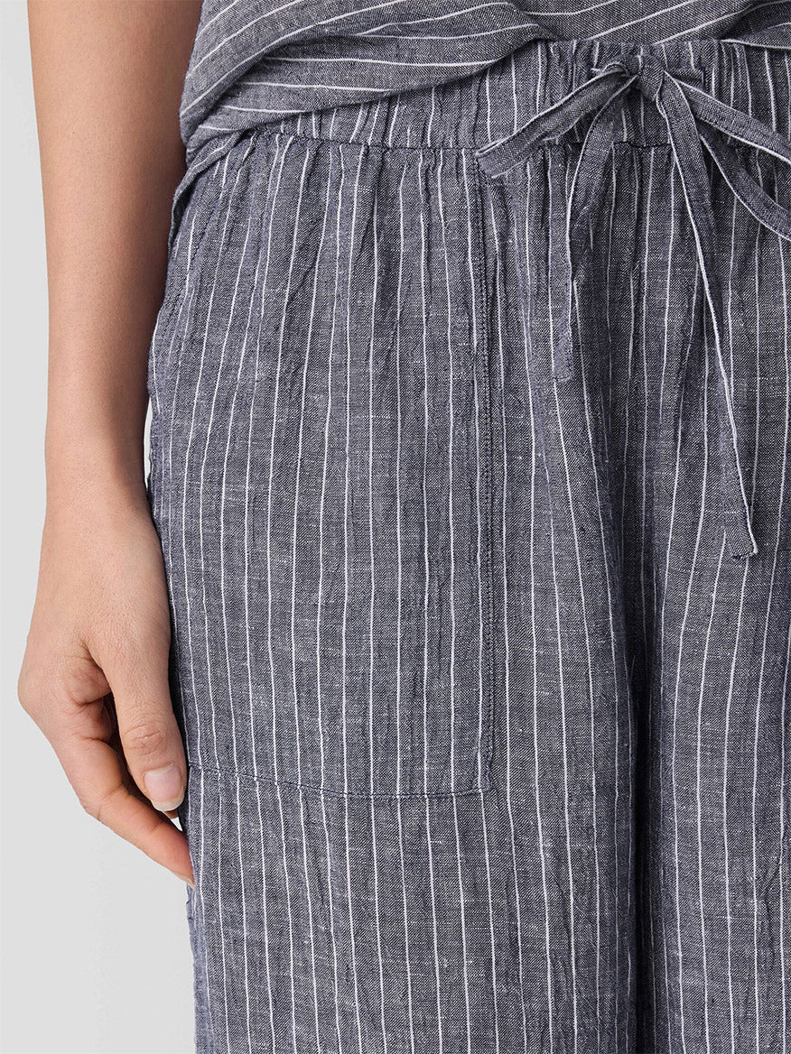 A close-up shows a person in Eileen Fisher Puckered Organic Linen Wide-Leg Pant in Black/White, featuring white vertical stripes, tie waist, and front pocket. The relaxed fit ensures comfort as their left hand rests at their side.