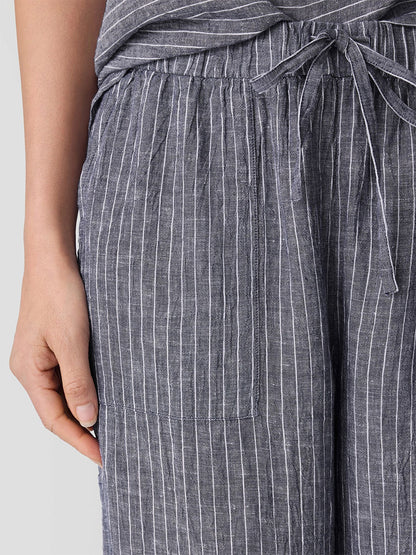 A close-up shows a person in Eileen Fisher Puckered Organic Linen Wide-Leg Pant in Black/White, featuring white vertical stripes, tie waist, and front pocket. The relaxed fit ensures comfort as their left hand rests at their side.