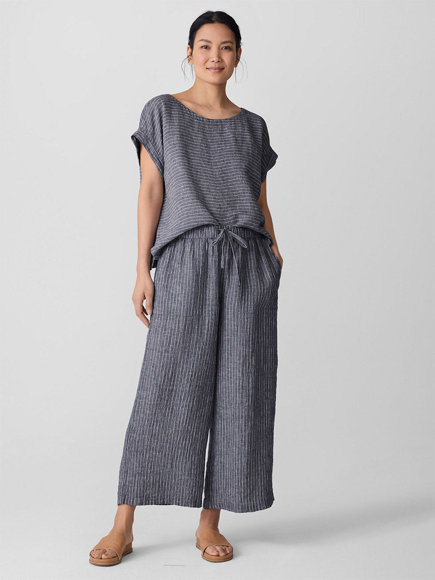 A woman stands facing forward wearing a loose-fitting, short-sleeve, gray striped top and Eileen Fisher Puckered Organic Linen Wide-Leg Pant in Black/White with sandals against a plain background.