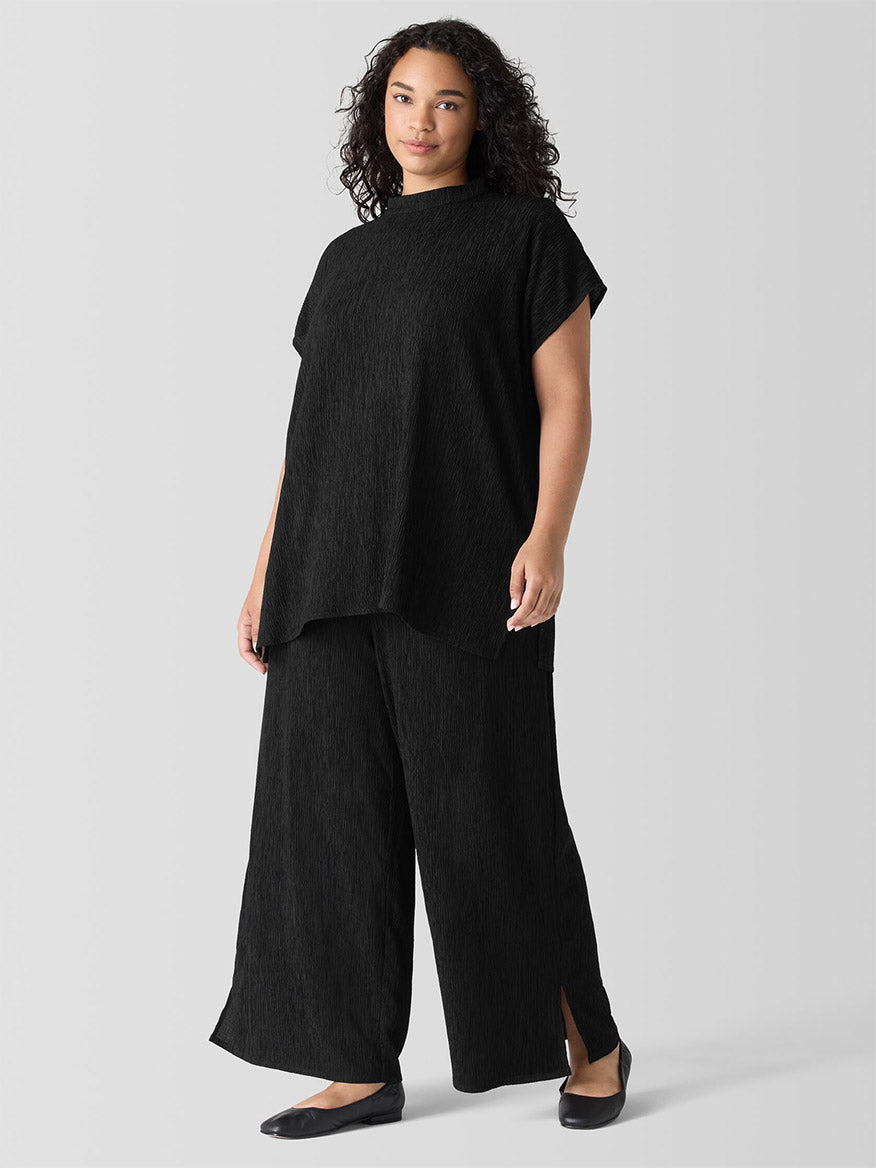 A woman with dark curly hair stands against a plain background, wearing a loose-fitting black top and the Eileen Fisher Woven Plissé Wide-Leg Pant in Black, paired with sleek black flats.