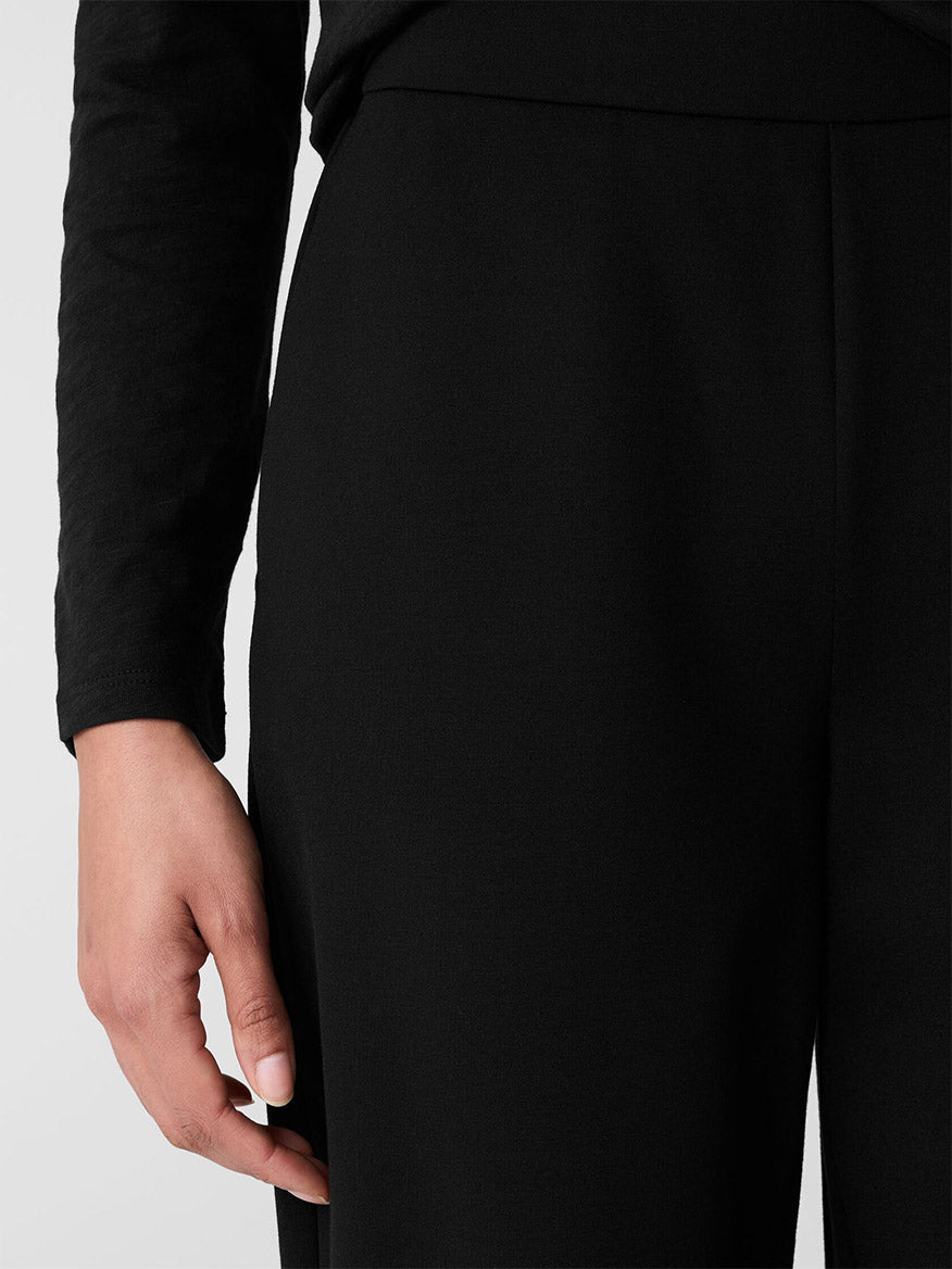 A person wearing a Fair Trade Certified, long-sleeved black shirt and Eileen Fisher Washable Flex Ponte Wide-Leg Pant in Black, with their left hand resting on their thigh, against a plain background.