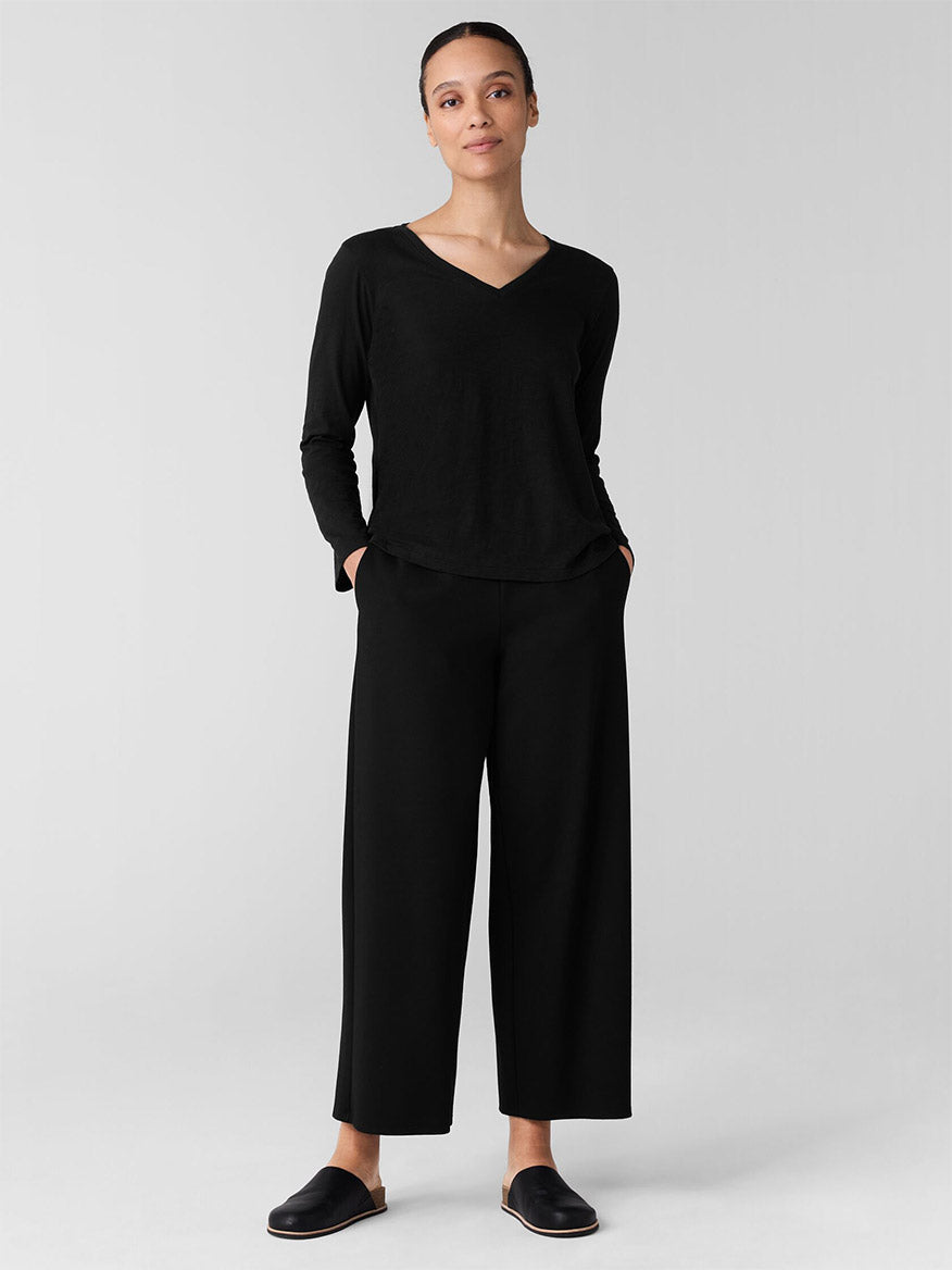 A person stands against a simple backdrop, wearing a black long-sleeve top, Eileen Fisher Washable Flex Ponte Wide-Leg Pant in Black, and black slip-on shoes.